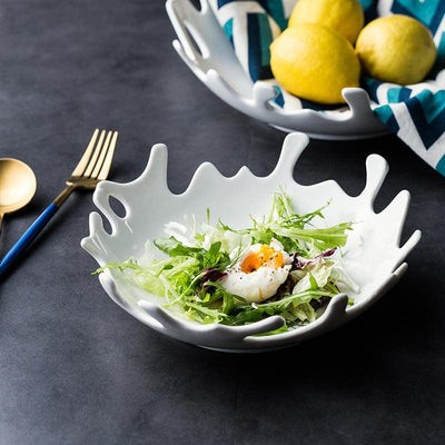 A white ceramic splash bowl with an irregular shape, containing a salad. A fork, spoon, and lemon are placed beside it on a dark countertop.