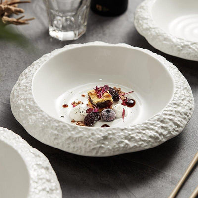 A white ceramic deep plate with a rock textured edge, displaying a portion of food on the plate, with other plates and cutlery in the background.