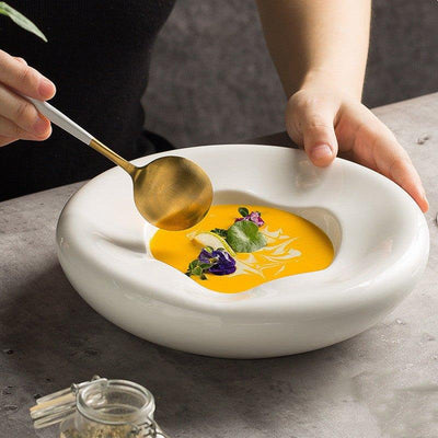 A white ceramic soup plate with an irregular shape, served with a vibrant orange soup garnished with green herbs and a touch of cream.