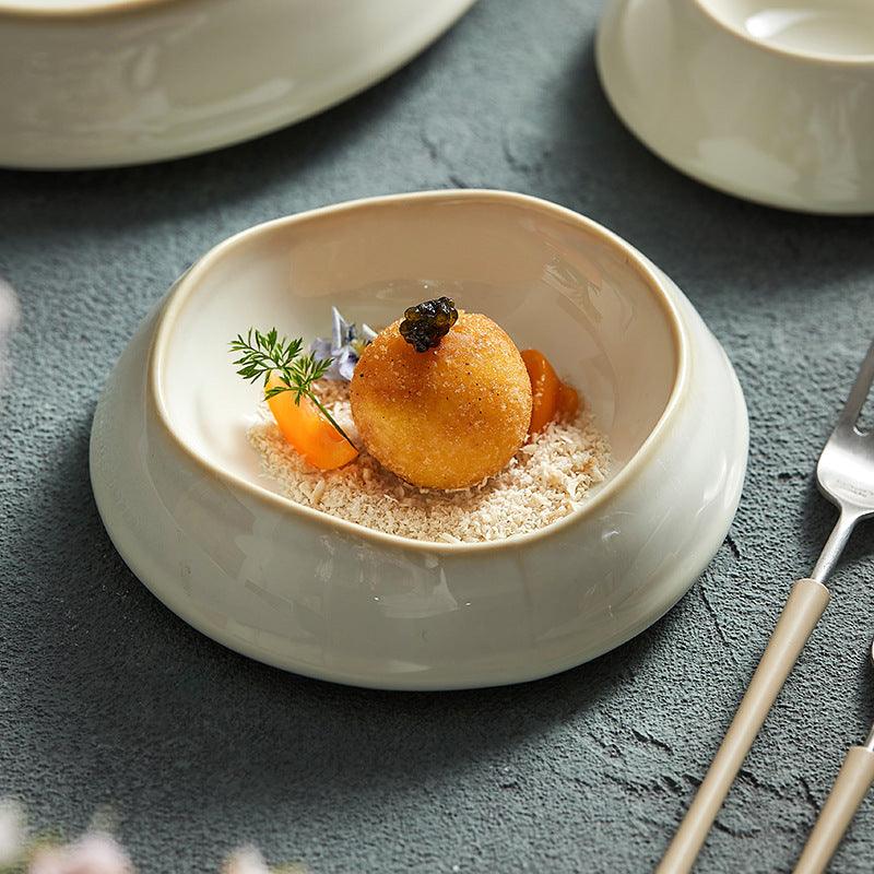Un plato gourmet presentado en un plato de cerámica de forma irregular. El plato es de color beige y presenta un elemento decorativo y un trozo de comida, posiblemente un postre.