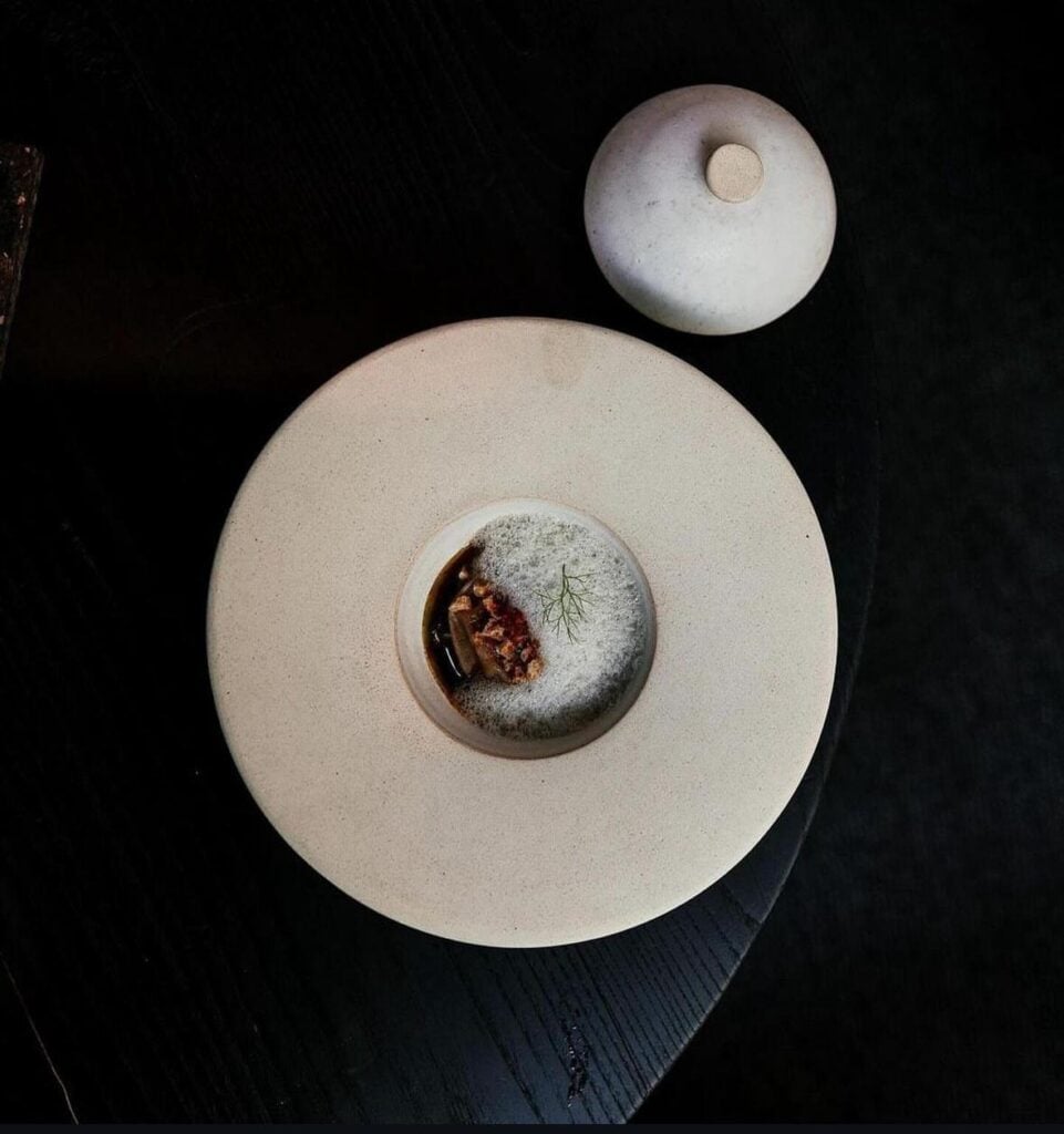 A white ceramic plate with a central depression for serving food, displayed alongside a smaller cloche functioning as a lid.
