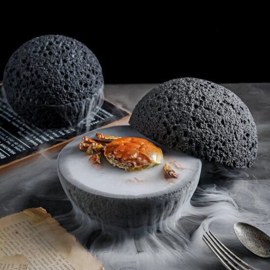 Close-up of a cooked crab centered in a split, volcanic-textured white bowl on a dark surface with theatrical white smoke.
